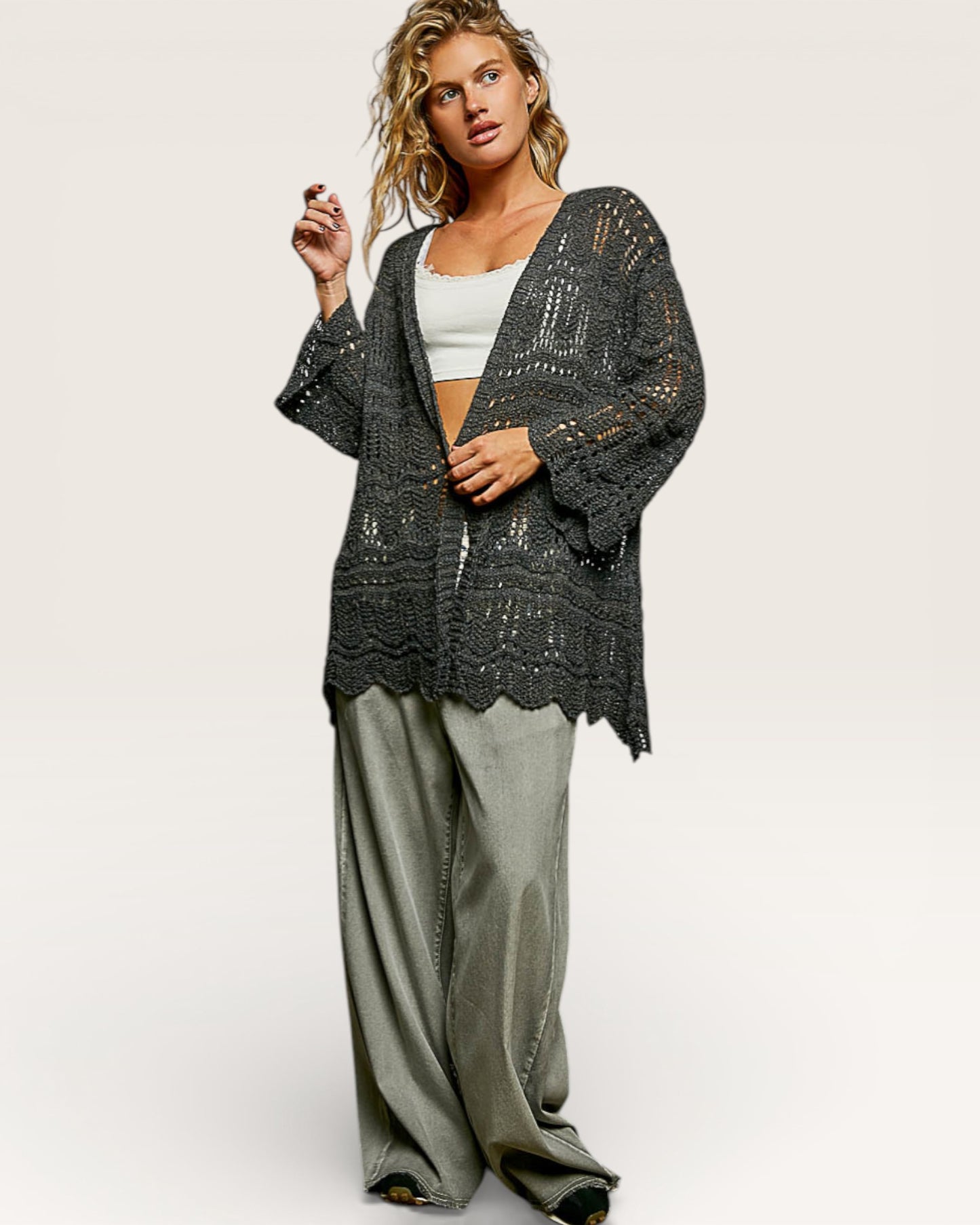Woman wearing a dark gray crochet cardigan over a white top and green pants on a light background