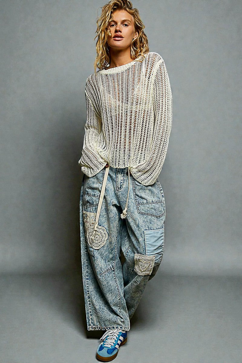 Person wearing a white knit sweater and blue jeans with patches on a gray background