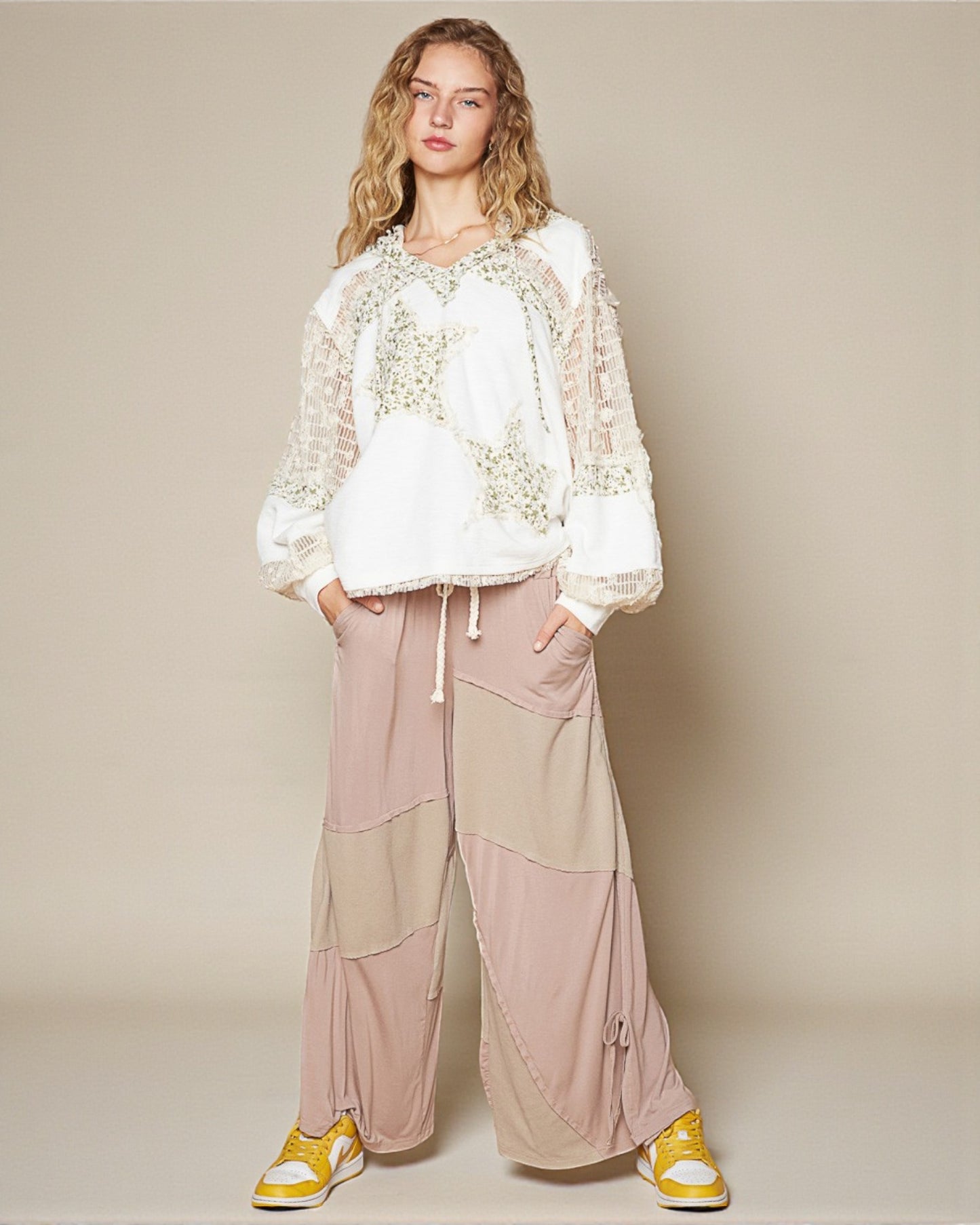 Woman wearing a white lace top and pink pants with yellow shoes on a beige background