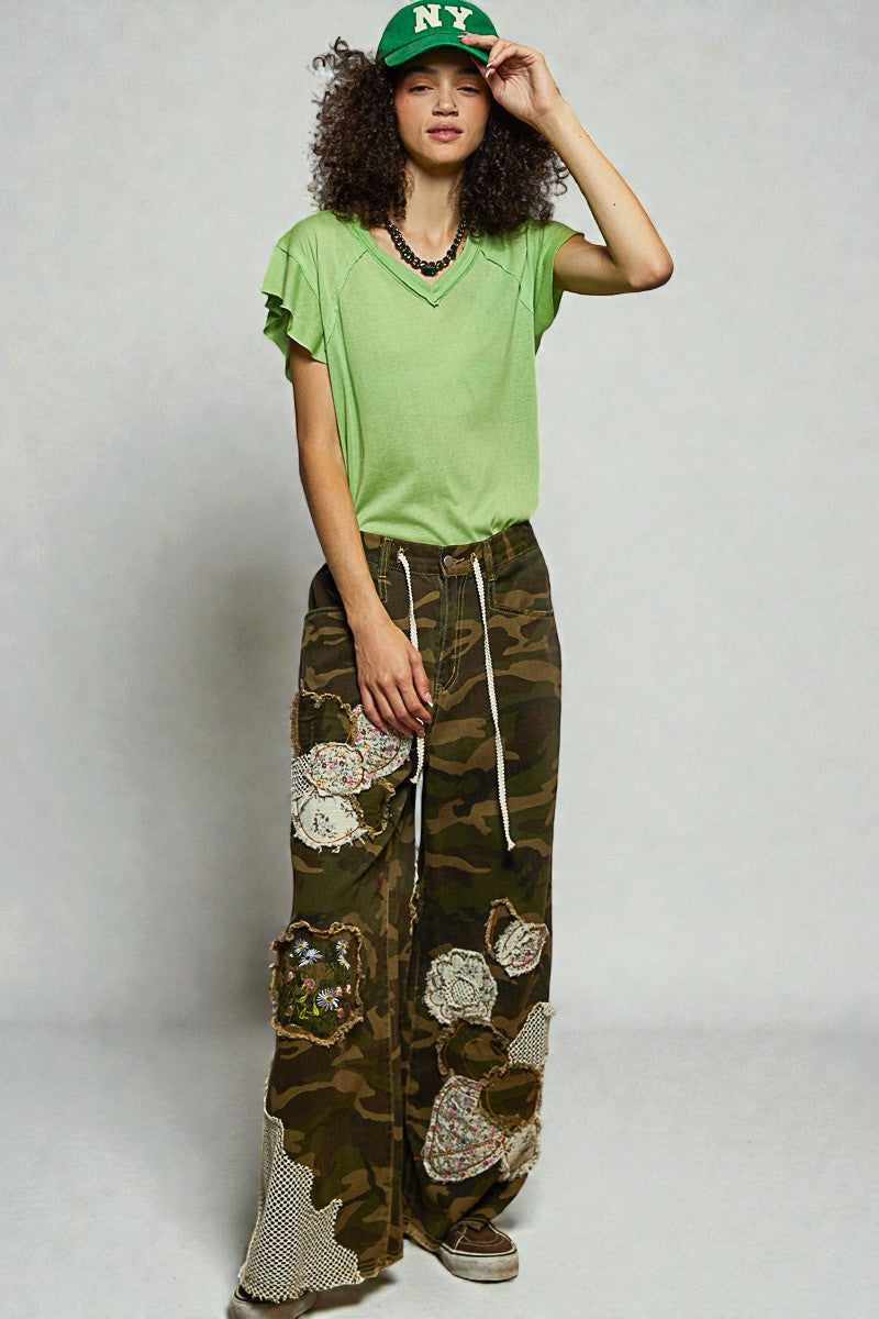 Person wearing a green t-shirt, camouflage pants with floral patches, and a green cap on a plain background