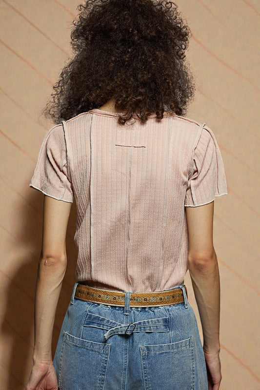 Person wearing a beige short-sleeve shirt and blue jeans with a patterned belt, standing against a beige wall.