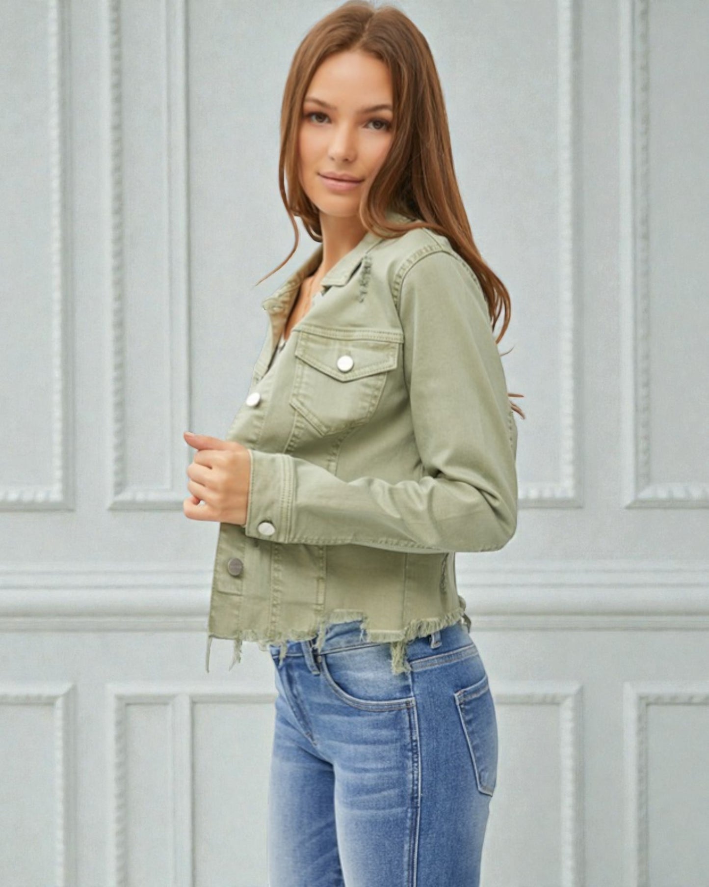 Woman wearing a green jacket and blue jeans against a light gray wall.