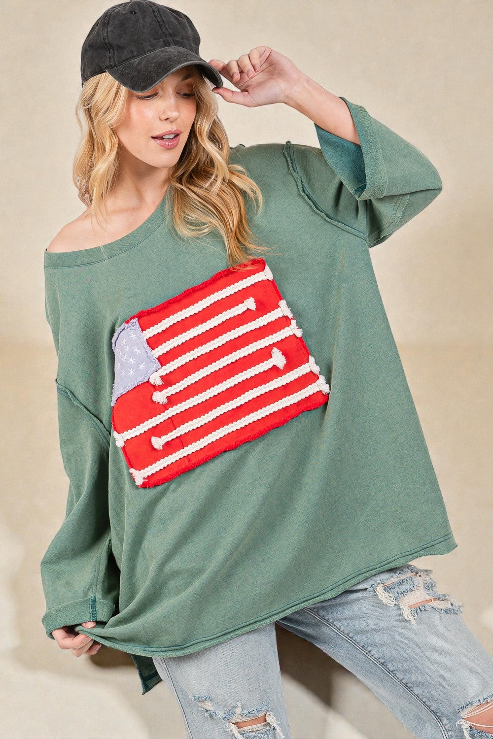 Woman wearing a green oversized sweater with a red and white graphic design, holding a black cap.