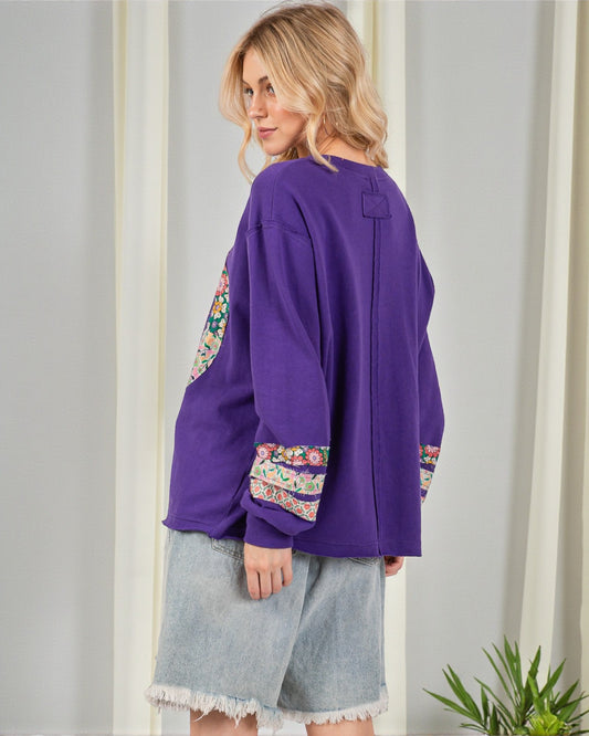 Woman wearing a purple top with floral cuffs and hem, standing against a white wall.