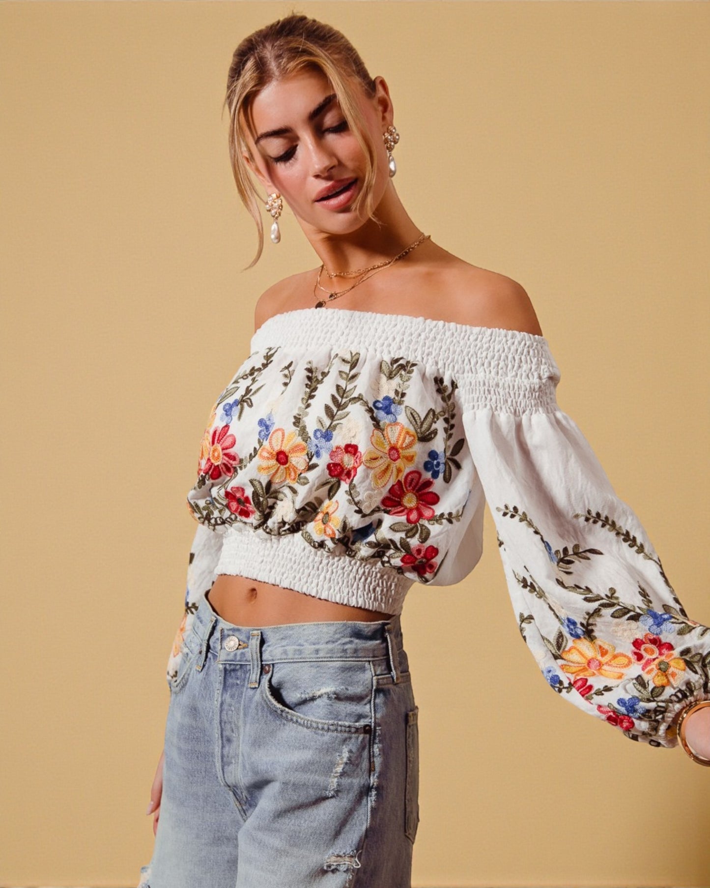 Woman wearing a white off-shoulder top with floral embroidery and light blue jeans on a beige background