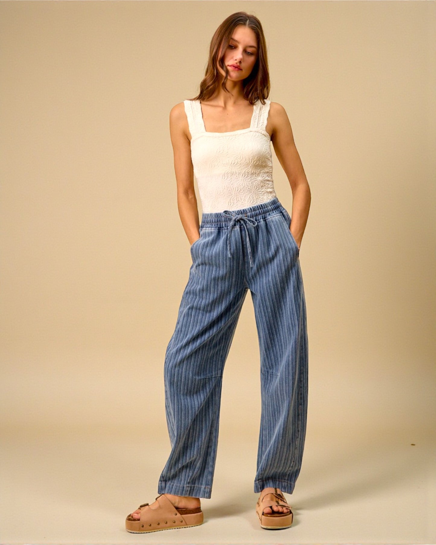 Woman wearing a white sleeveless top and blue striped pants against a beige background