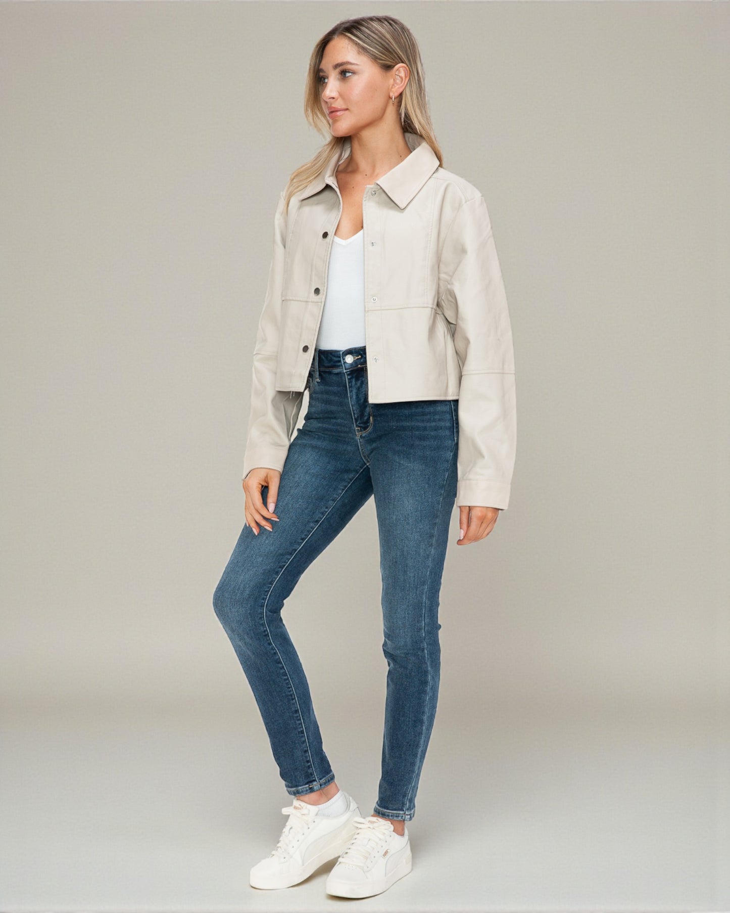 Woman wearing a beige jacket, white shirt, blue jeans, and white sneakers on a plain background