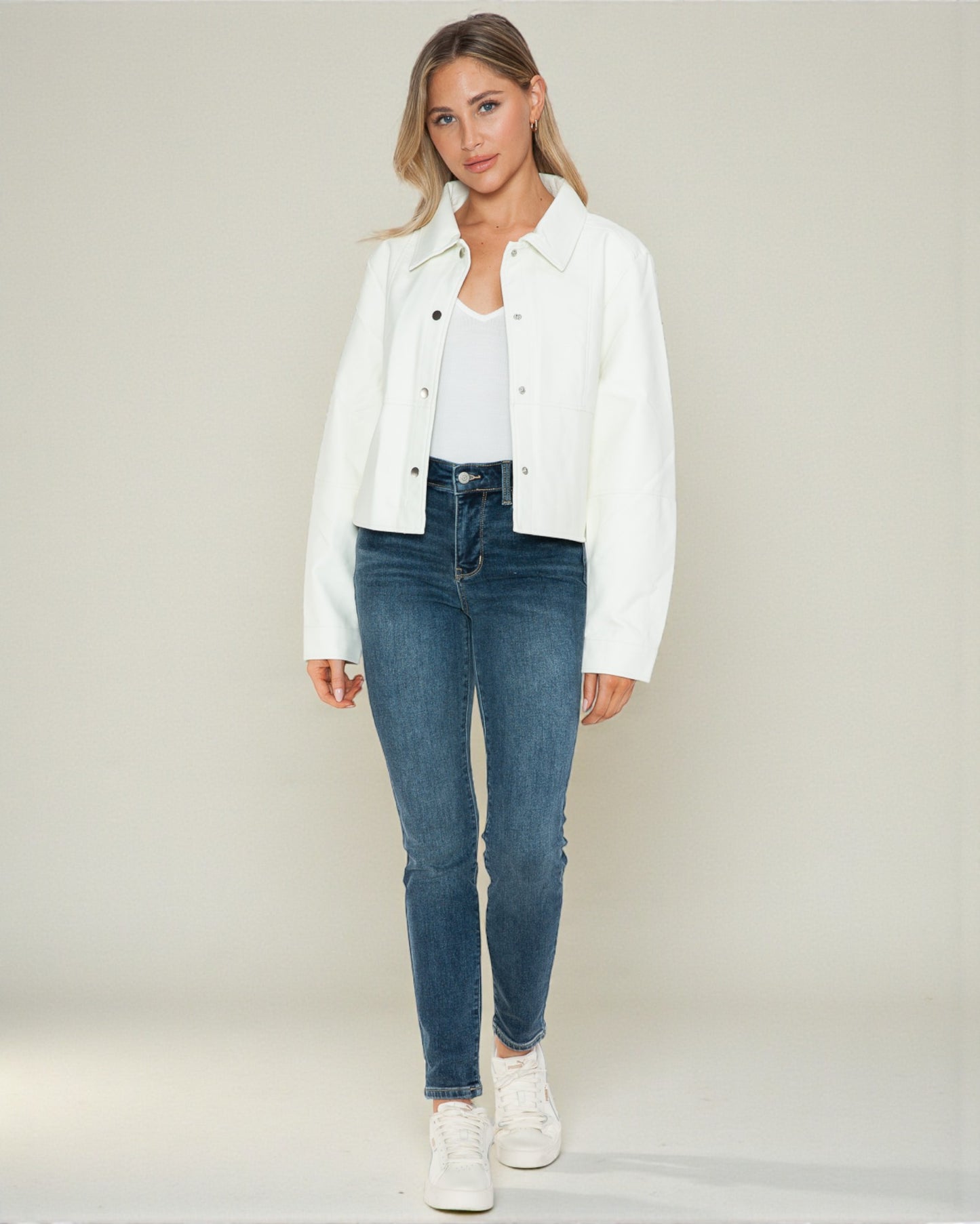 Woman wearing a white jacket, white shirt, blue jeans, and white sneakers on a plain background