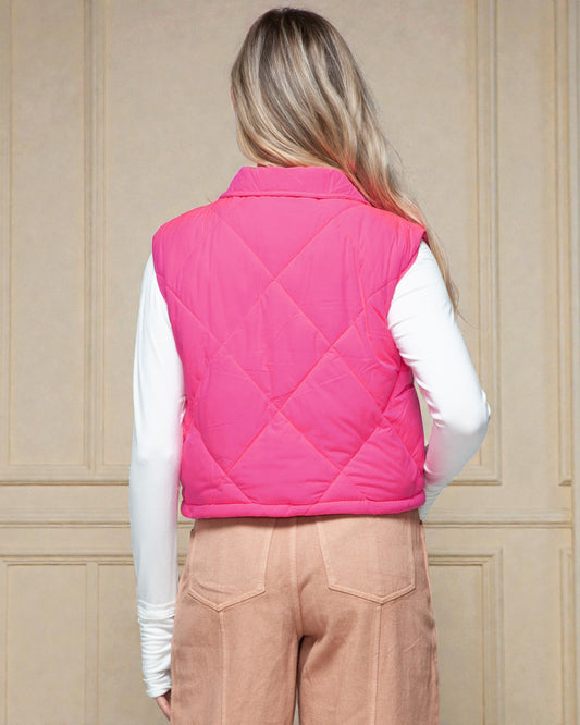 Person wearing a pink quilted vest over a white shirt and beige pants against a beige paneled wall.