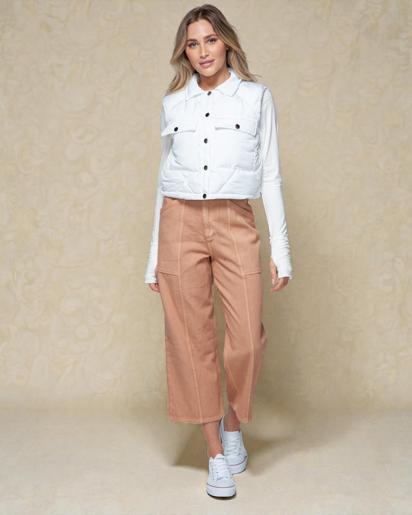 Woman wearing a white quilted jacket, white shirt, tan pants, and white sneakers on a beige background