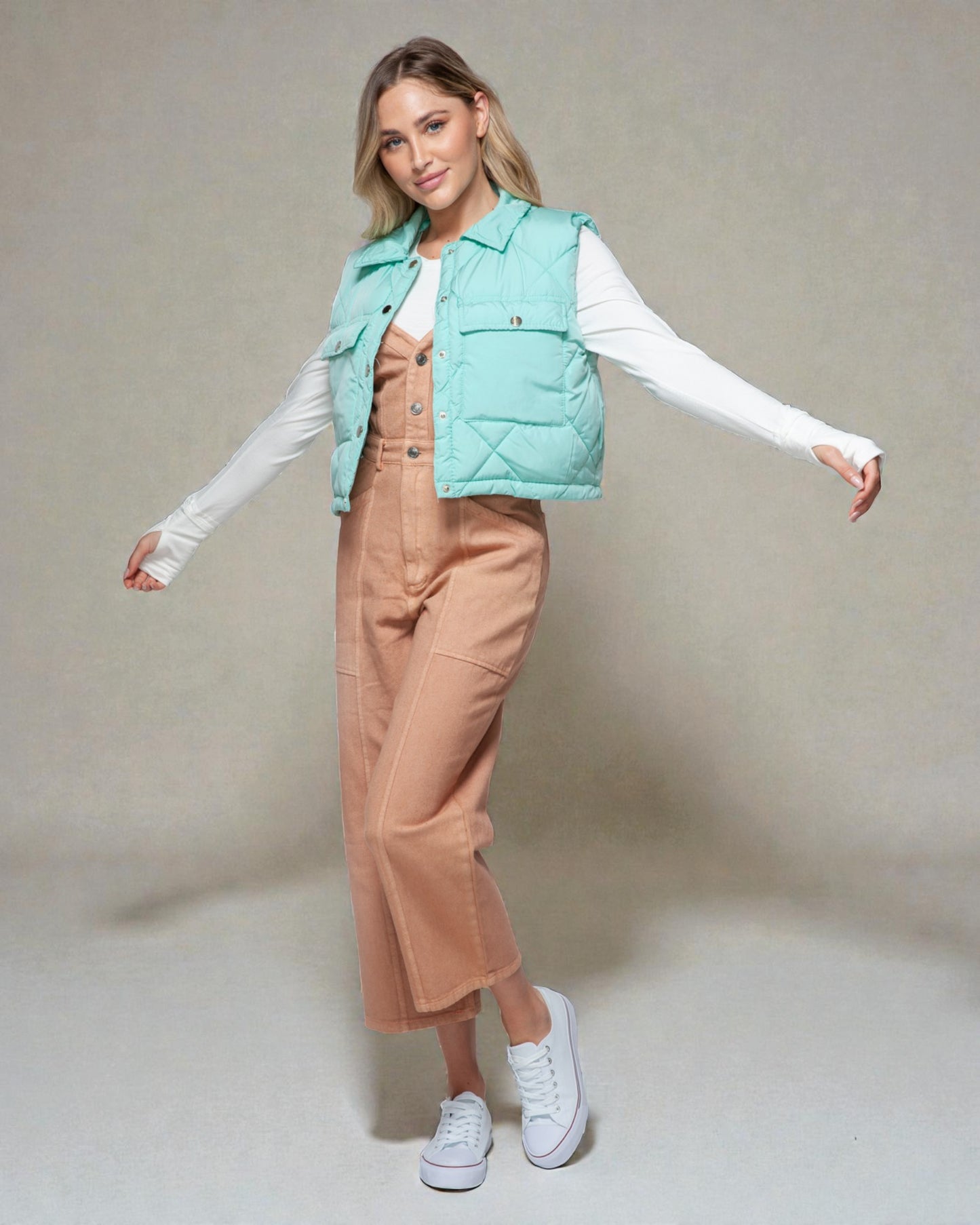 Woman wearing a light blue puffer vest over a white shirt and beige pants on a plain background