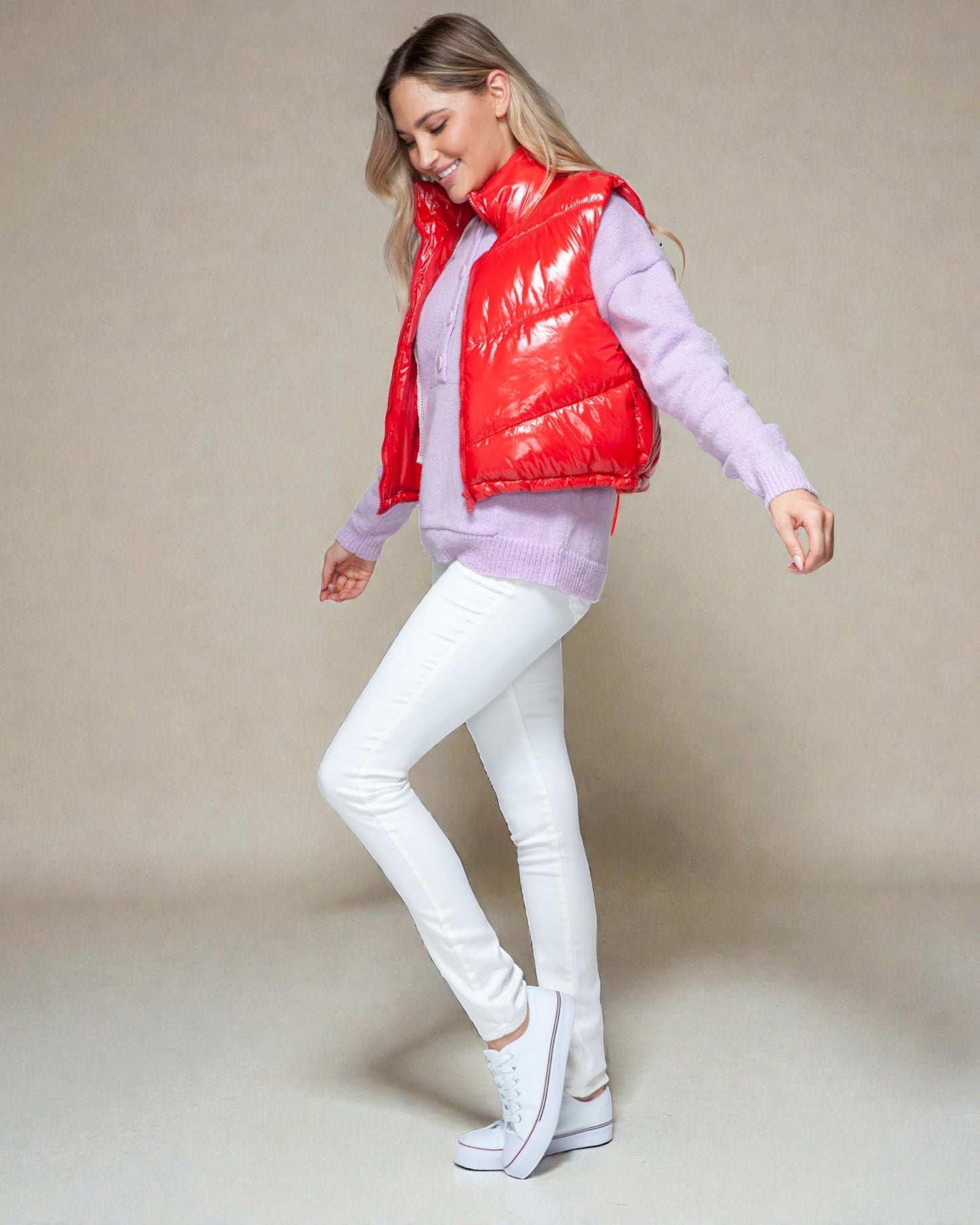 Woman wearing a red puffer vest, light purple sweater, white pants, and white sneakers on a beige background