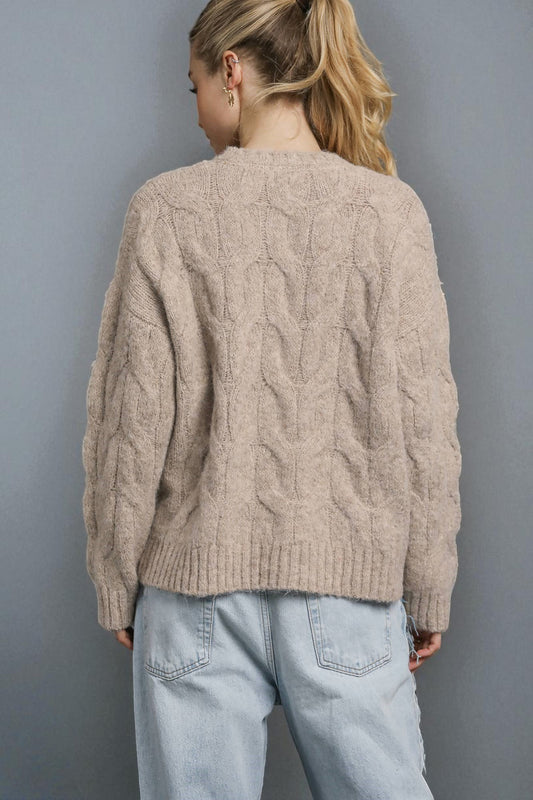 Person wearing a beige cable knit sweater and light blue jeans against a gray background