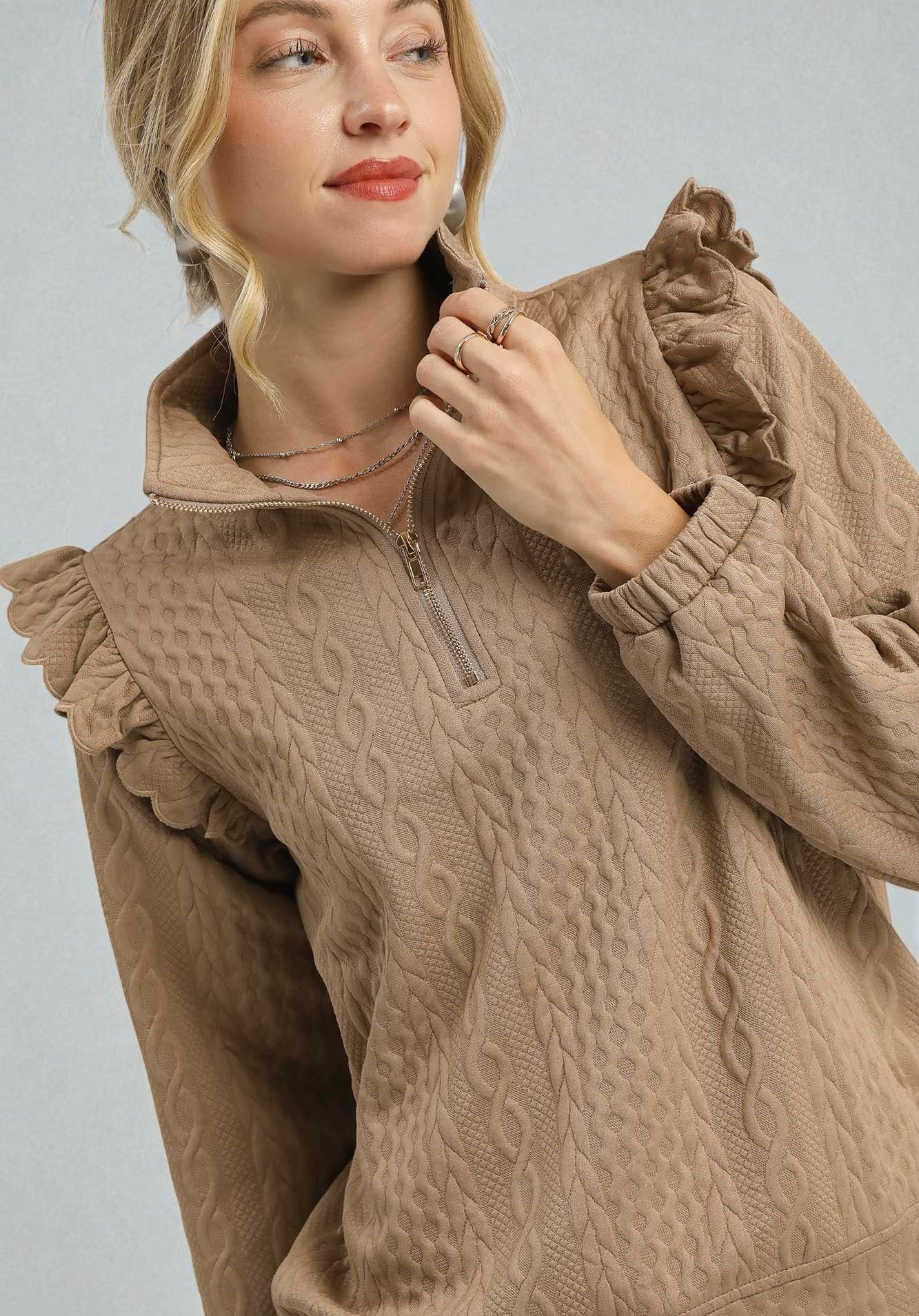 Woman wearing a beige cable knit sweater with ruffled details on a light gray background