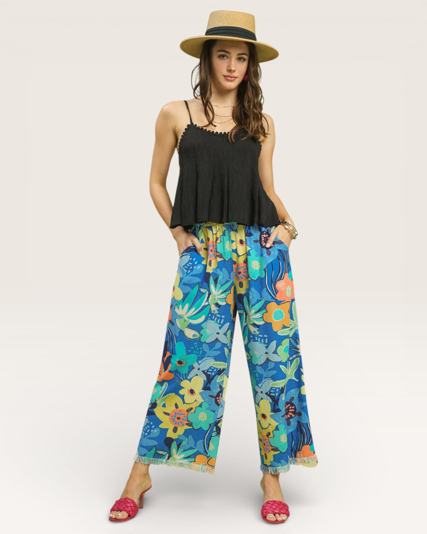 Woman wearing a black top and colorful floral pants with a straw hat on a white background