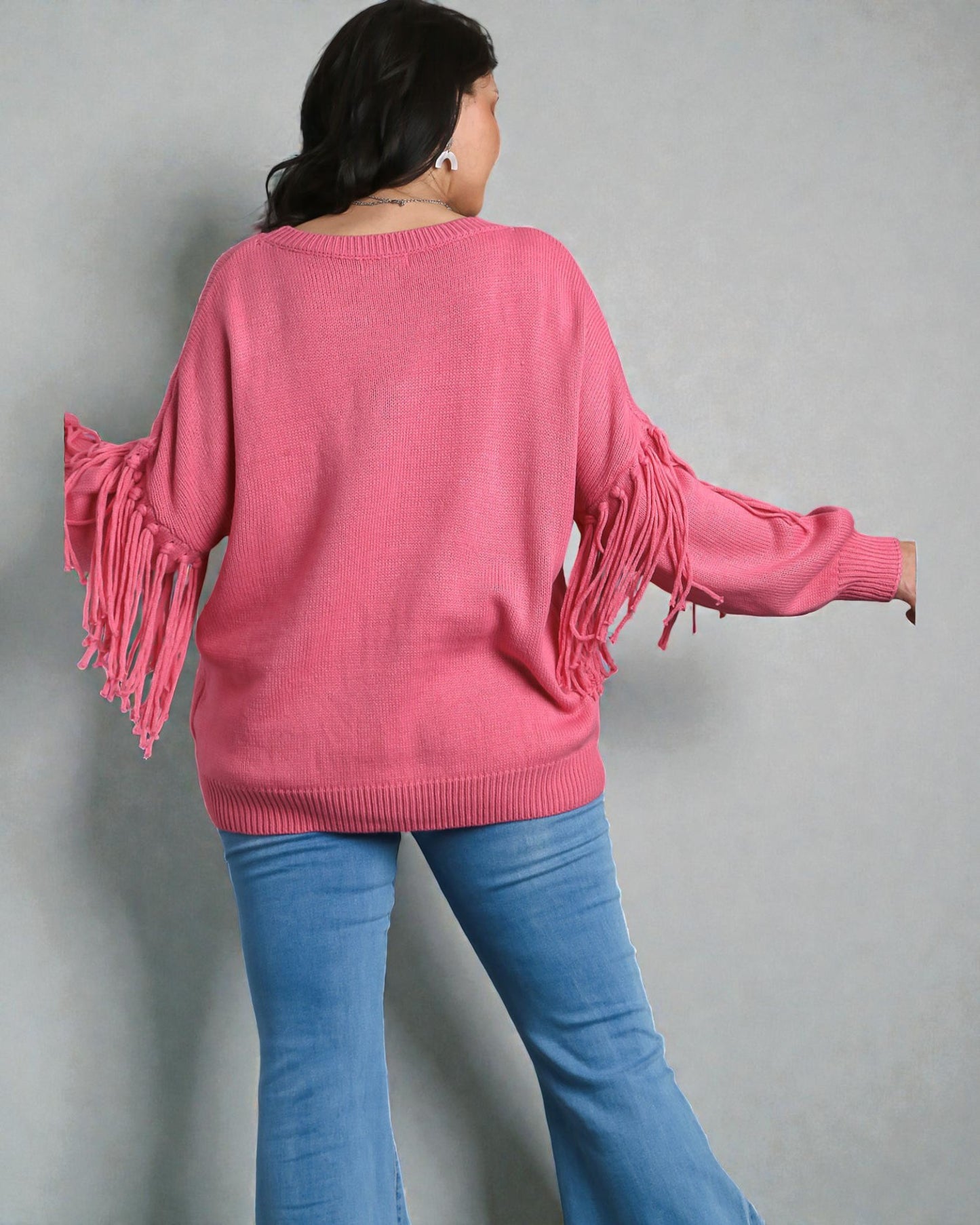 Person wearing a pink sweater with fringe details and blue jeans against a gray background