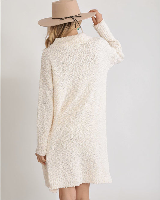 Person wearing a cream-colored sweater dress and beige hat on a plain background