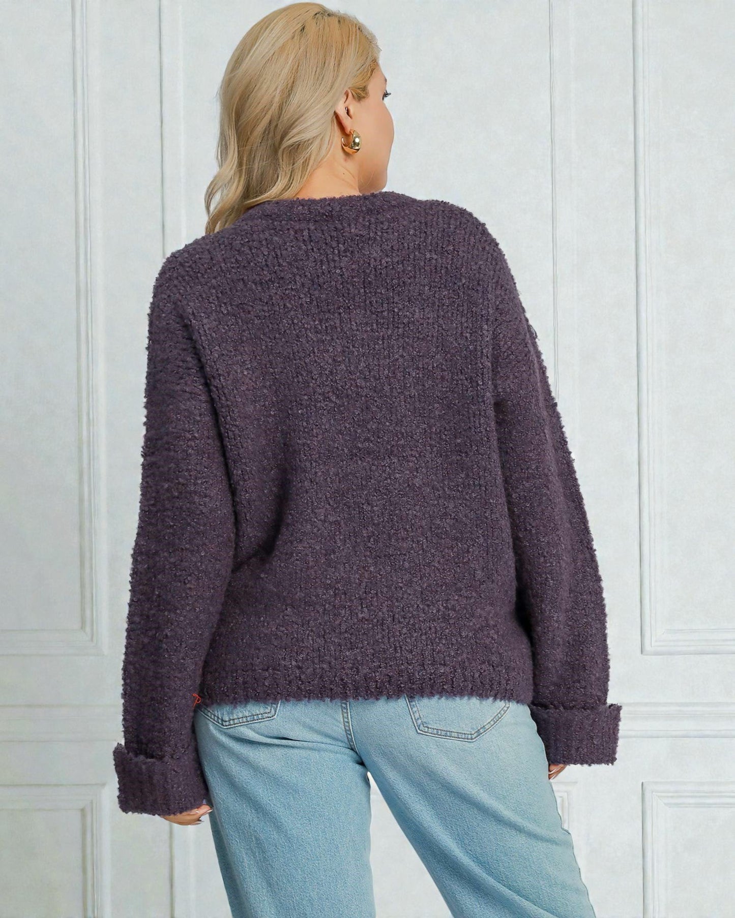 Person wearing a purple textured sweater and light blue jeans against a white paneled wall.