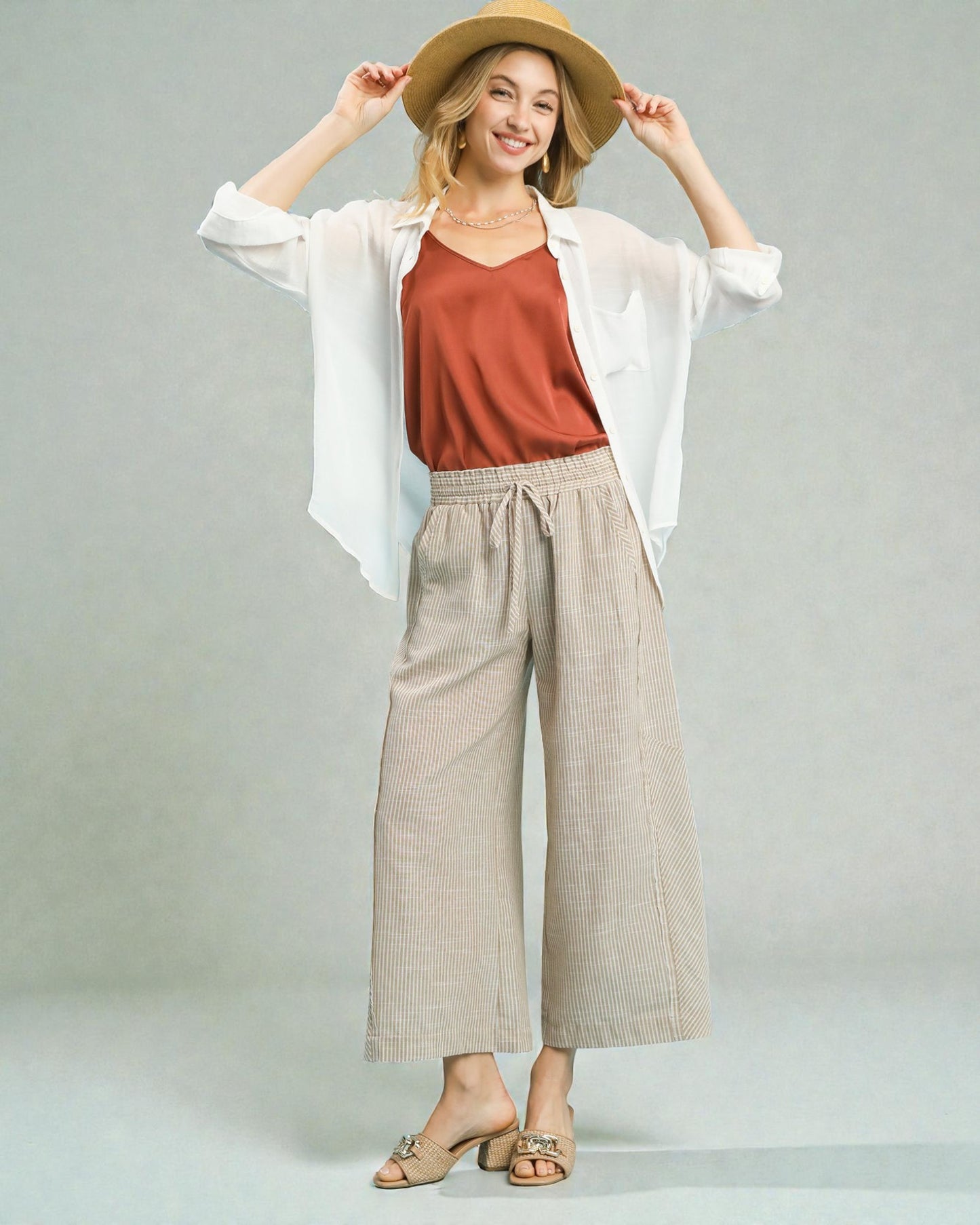 Woman wearing a white shirt, rust-colored top, beige wide-leg pants, and a straw hat on a gray background