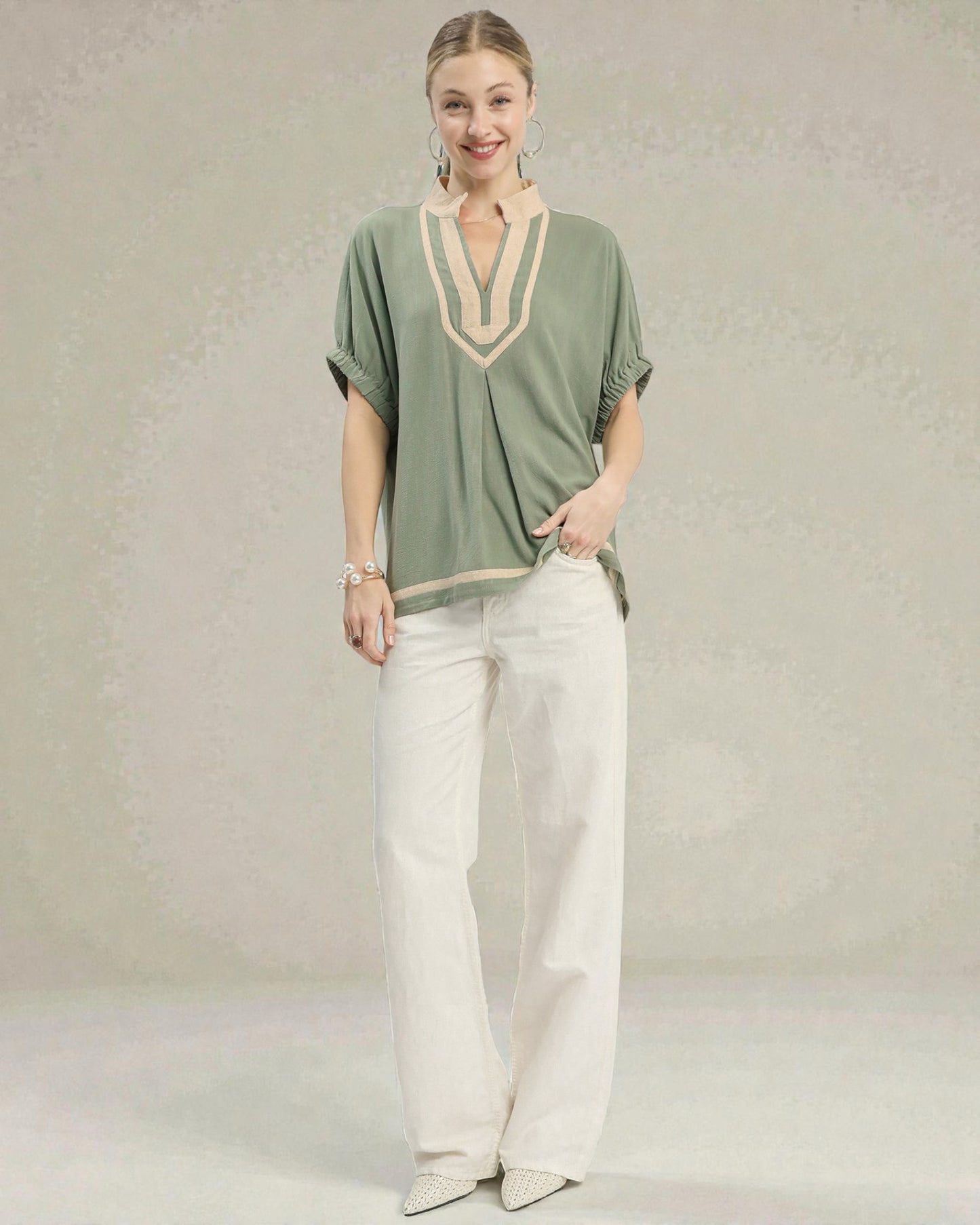 Woman wearing a green top with beige accents and white pants on a plain background
