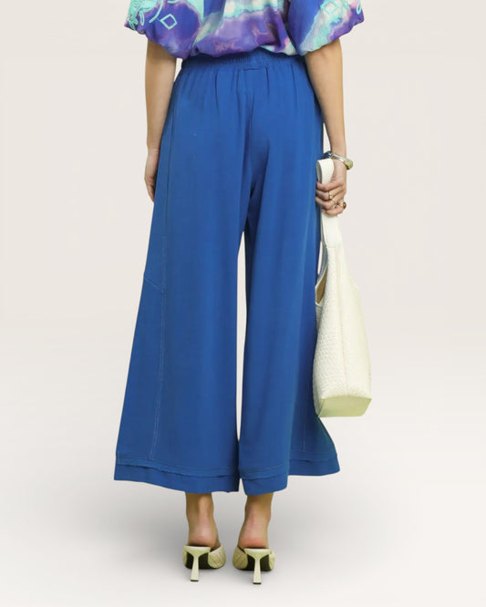 Person wearing blue wide-leg pants and a colorful top, holding a white bag on a light background