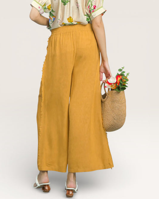 Person wearing mustard yellow wide-leg pants holding a woven bag with flowers on a plain background
