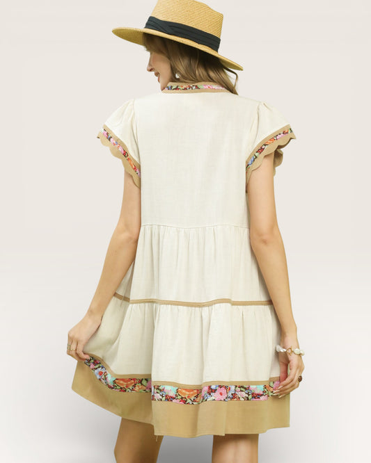 Woman wearing a cream-colored dress with floral embroidery and a straw hat on a plain background
