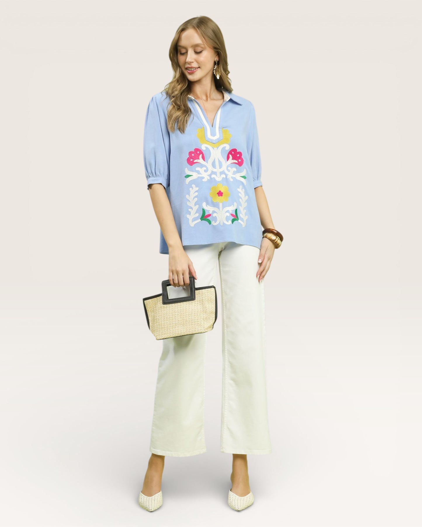 Woman wearing a light blue embroidered blouse with white pants, holding a straw bag on a plain background