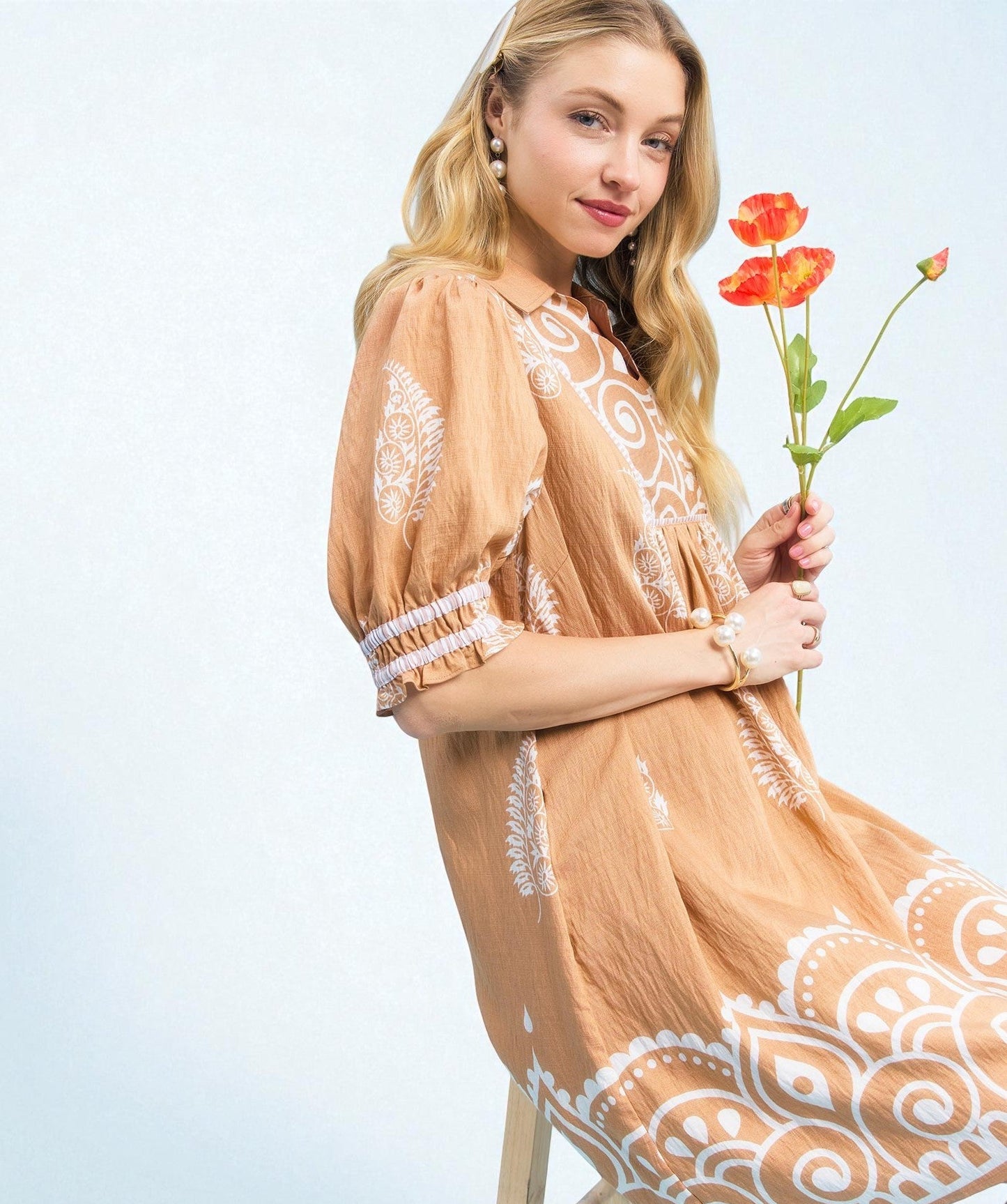 Woman in a beige dress with white lace details holding red flowers against a light blue background