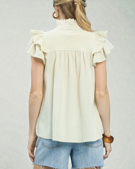 Woman wearing a cream blouse with ruffled shoulders and denim shorts on a plain background