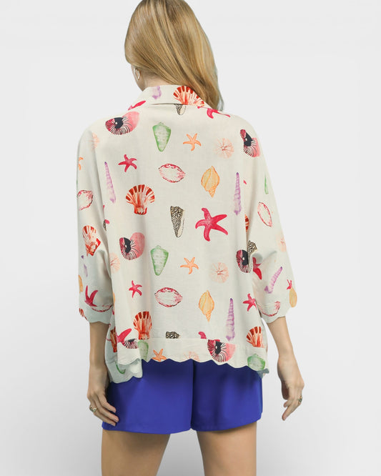 Woman wearing a colorful shell-patterned blouse with blue shorts on a white background