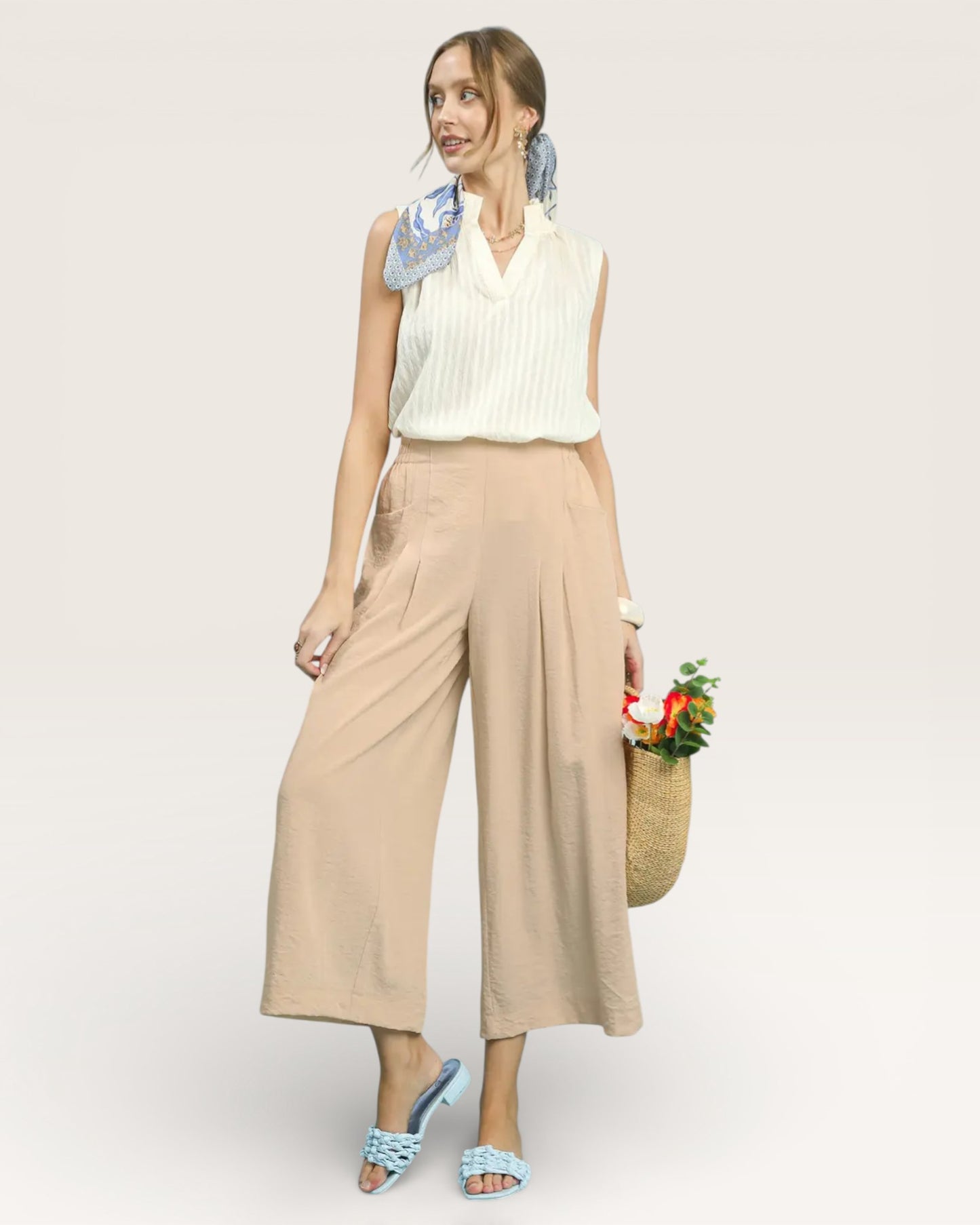 Woman wearing a white sleeveless top and beige wide-leg pants, holding flowers and a basket, on a light gray background.
