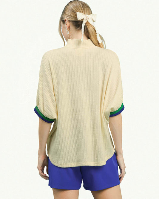 Person wearing a beige ribbed sweater with green and blue trim and blue shorts on a white background