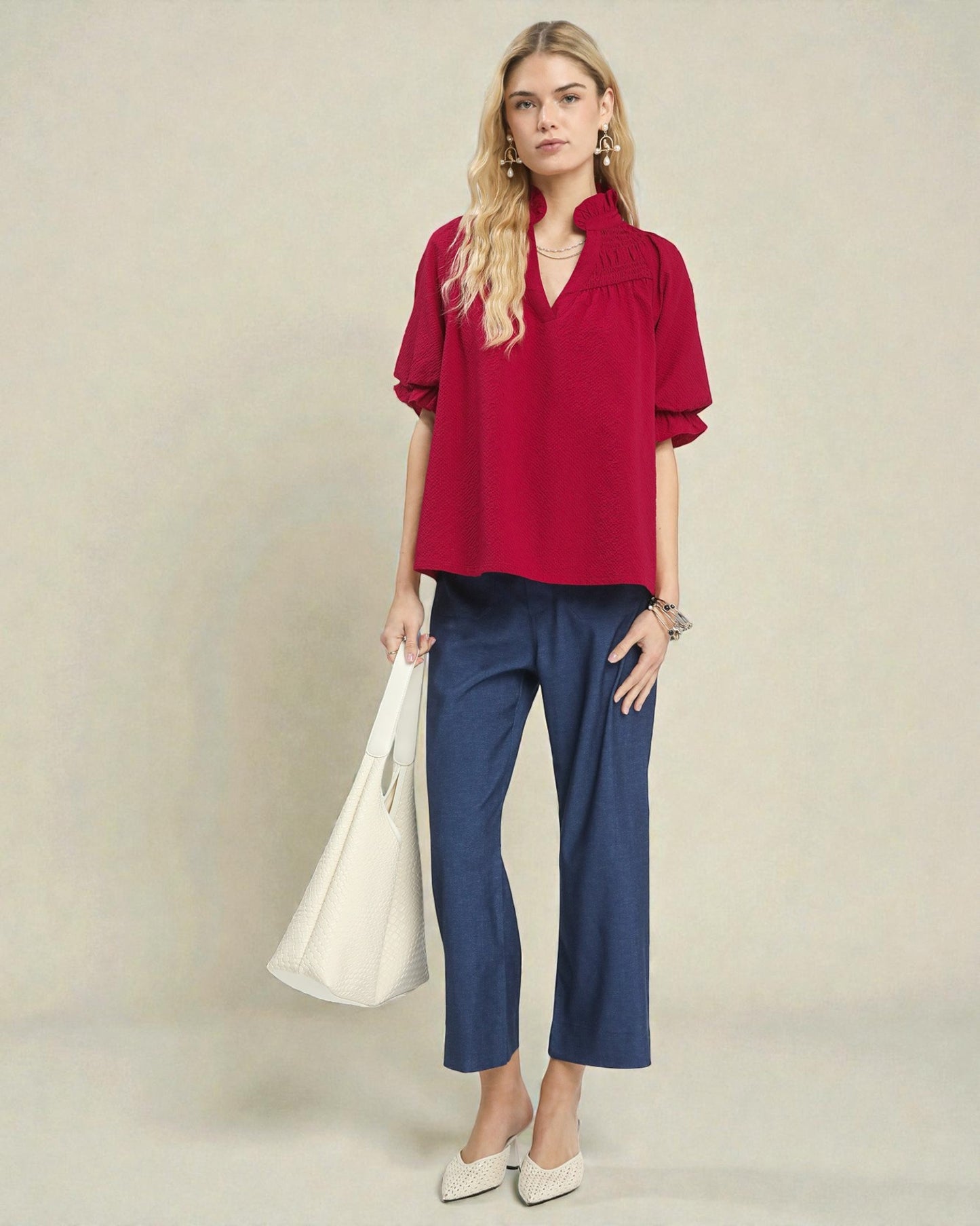 Woman wearing a red blouse and blue pants holding a white bag against a beige background