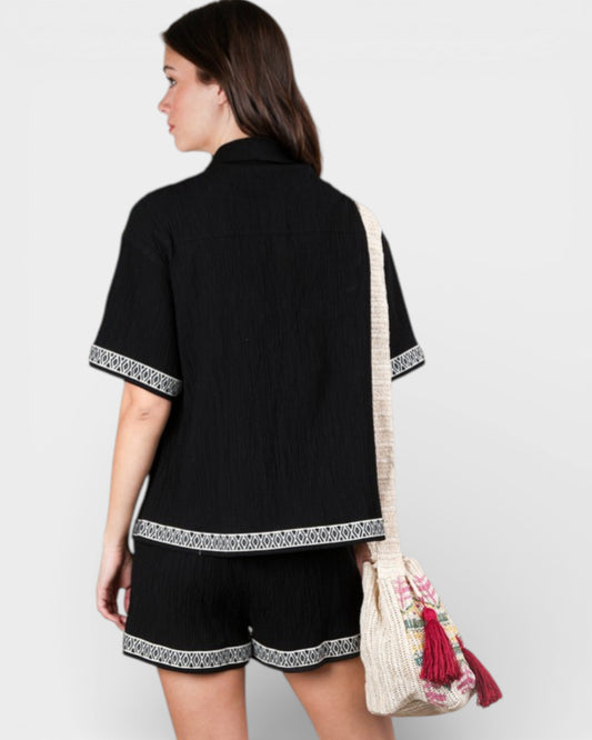 Woman wearing a black outfit with white patterns, holding a colorful bag on a plain background