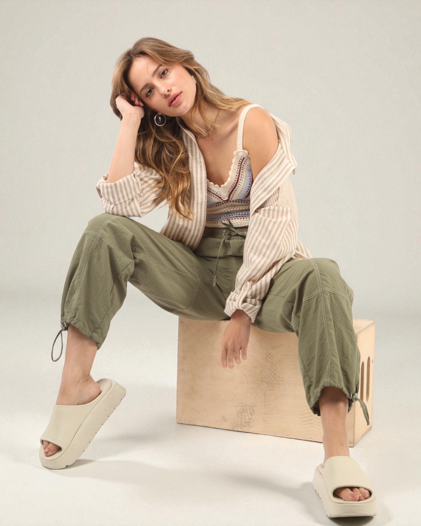 Woman sitting on a wooden block wearing a striped top and green pants against a plain background