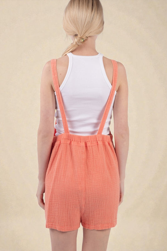 Person wearing a white tank top and coral checkered shorts with suspenders on a beige background