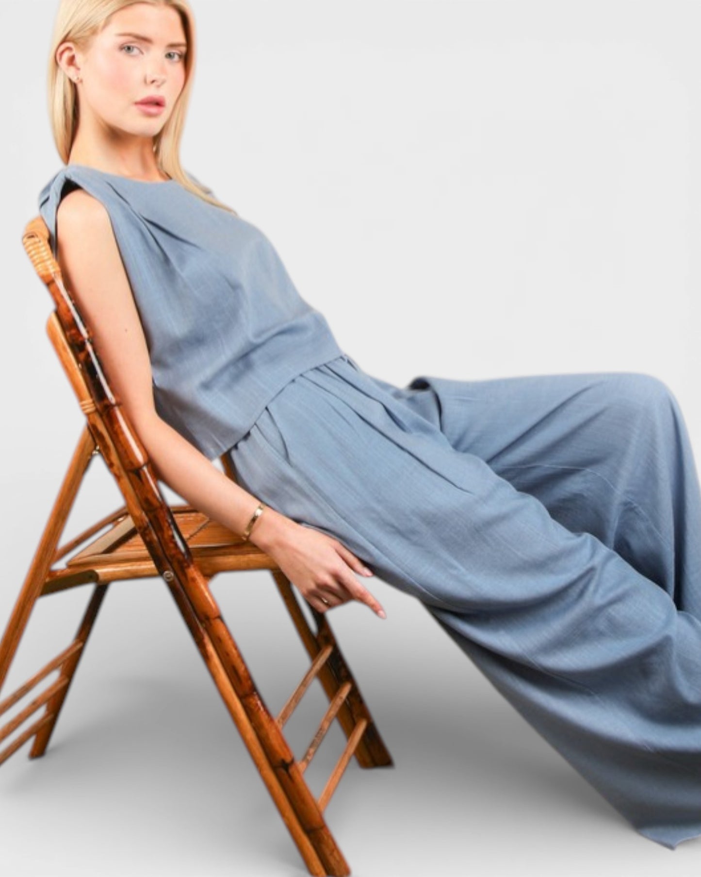 Woman wearing a blue jumpsuit sitting on a wooden chair against a light gray background