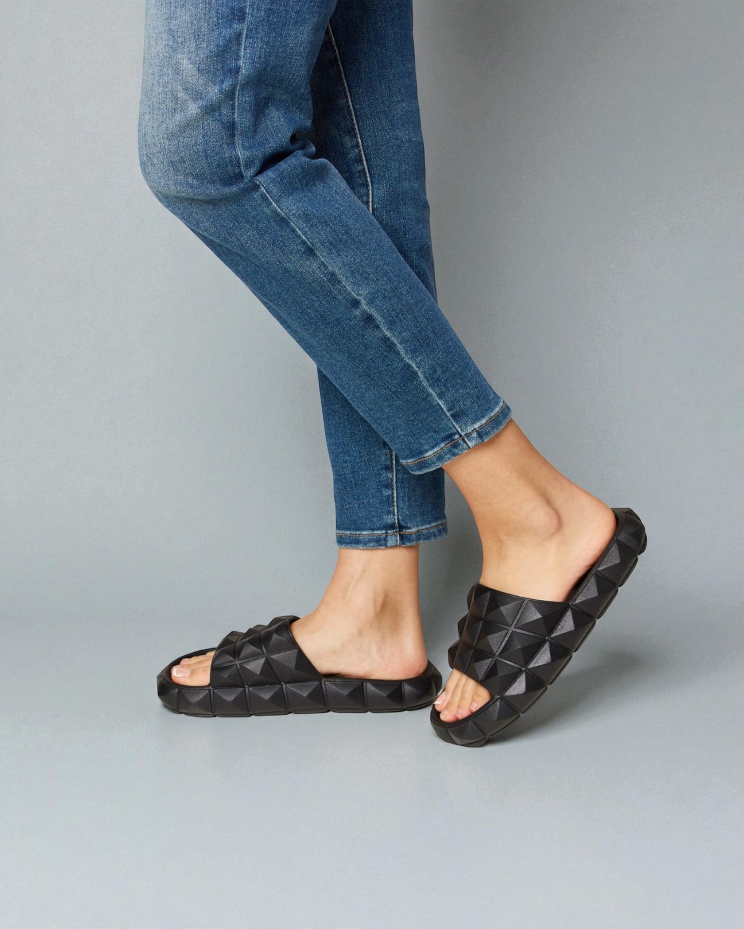 Person wearing black quilted sandals and blue jeans on a gray background