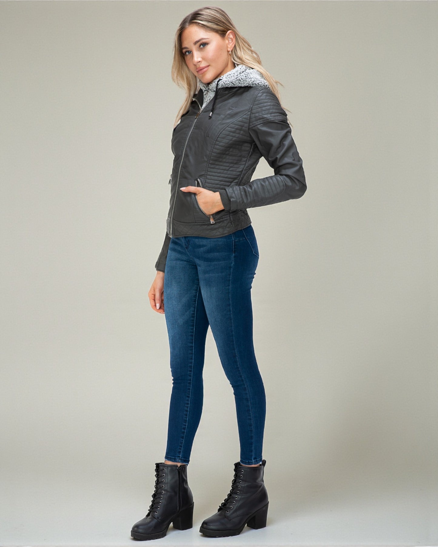 Woman wearing a gray jacket with a hood, blue jeans, and black boots on a plain background