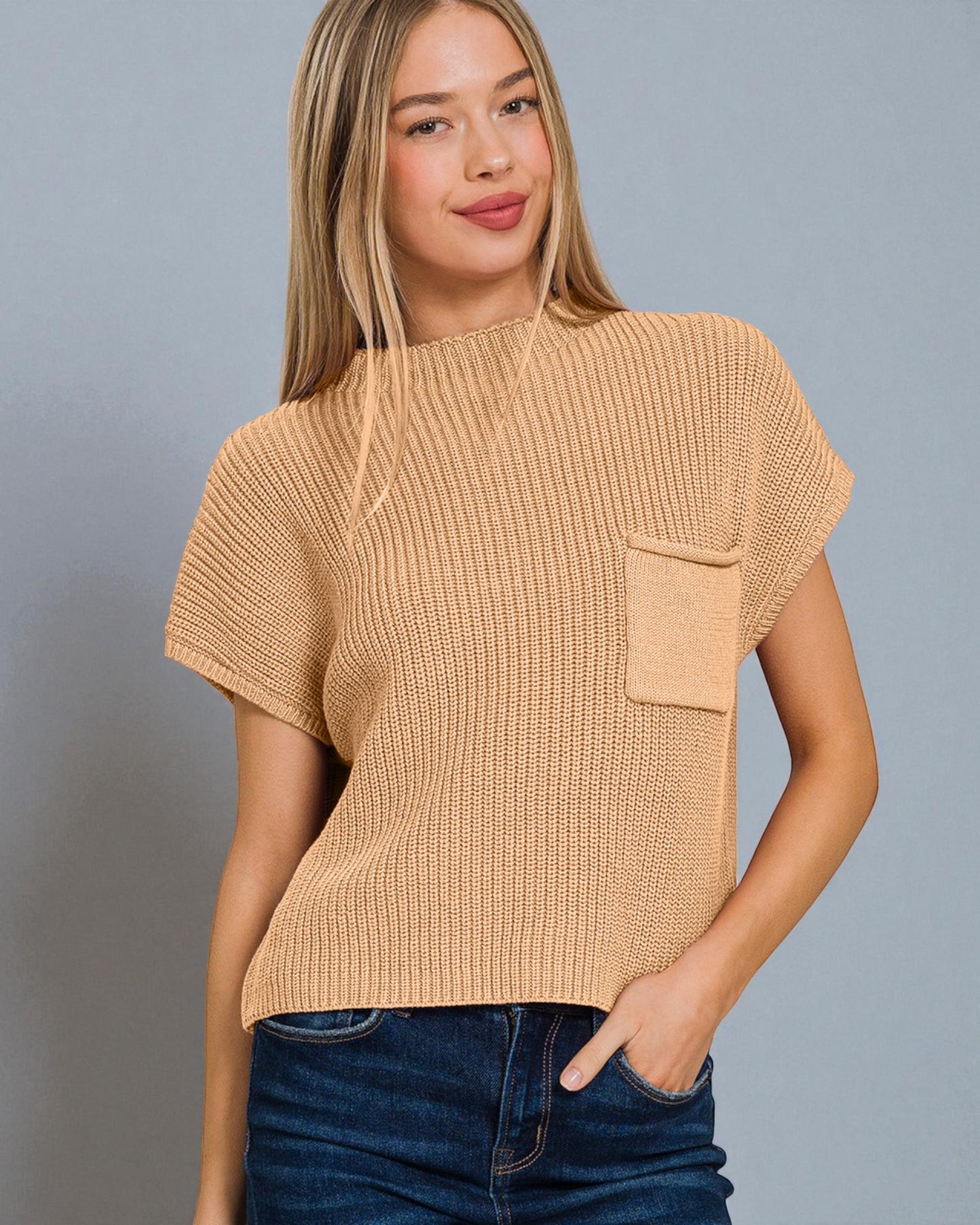 Woman wearing a beige knitted top with a pocket against a gray background