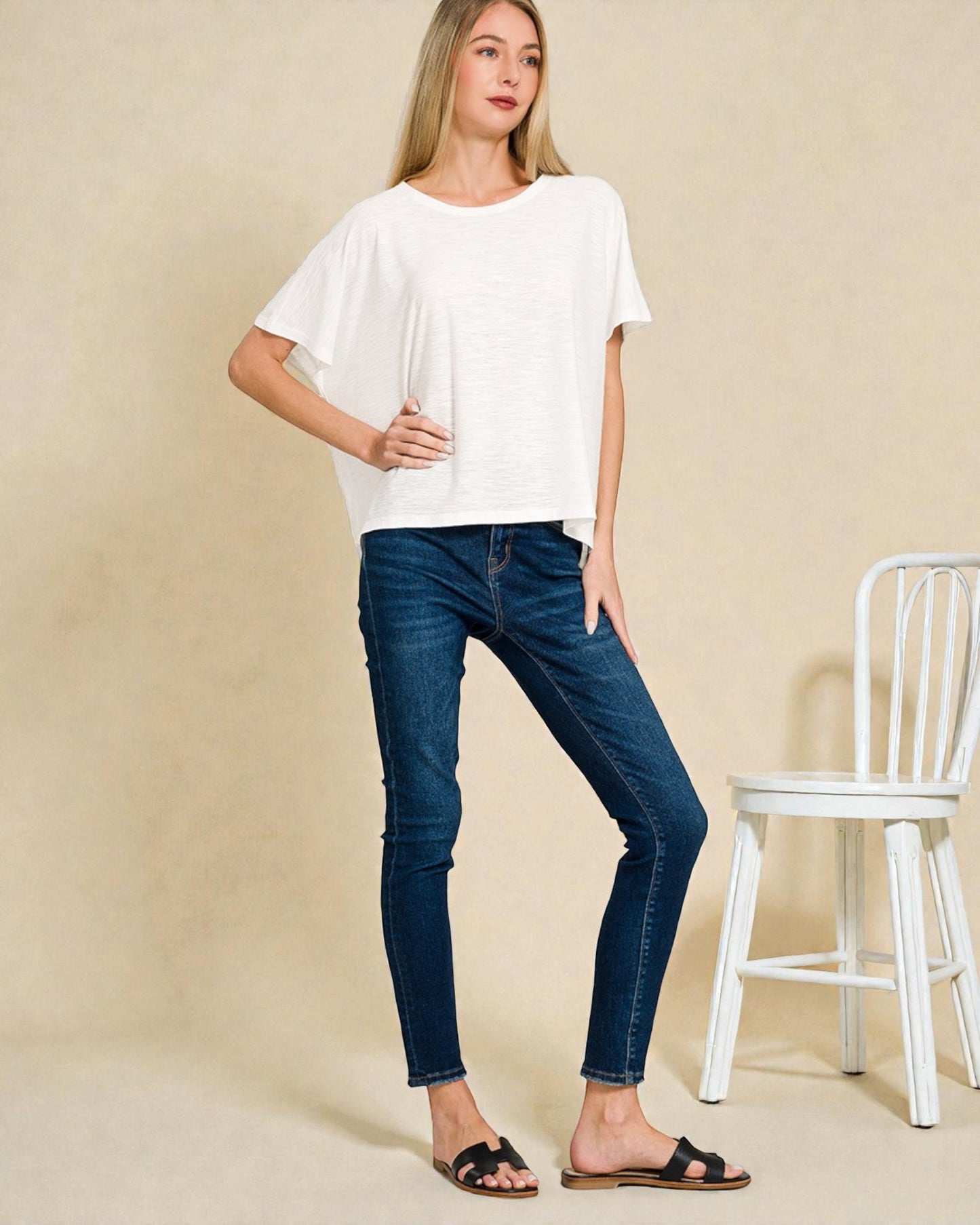 Woman wearing a white t-shirt and blue jeans standing against a beige background with a white chair.