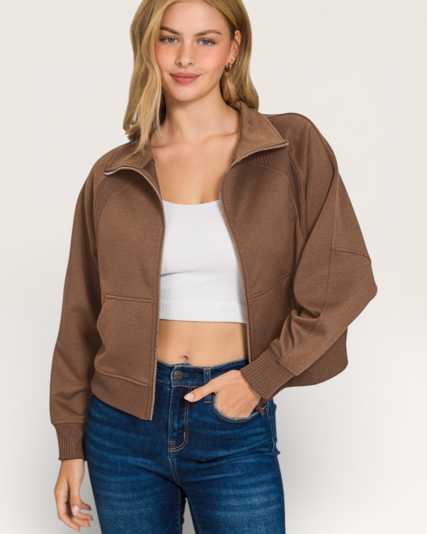 Woman wearing a brown jacket, white top, and blue jeans on a plain background