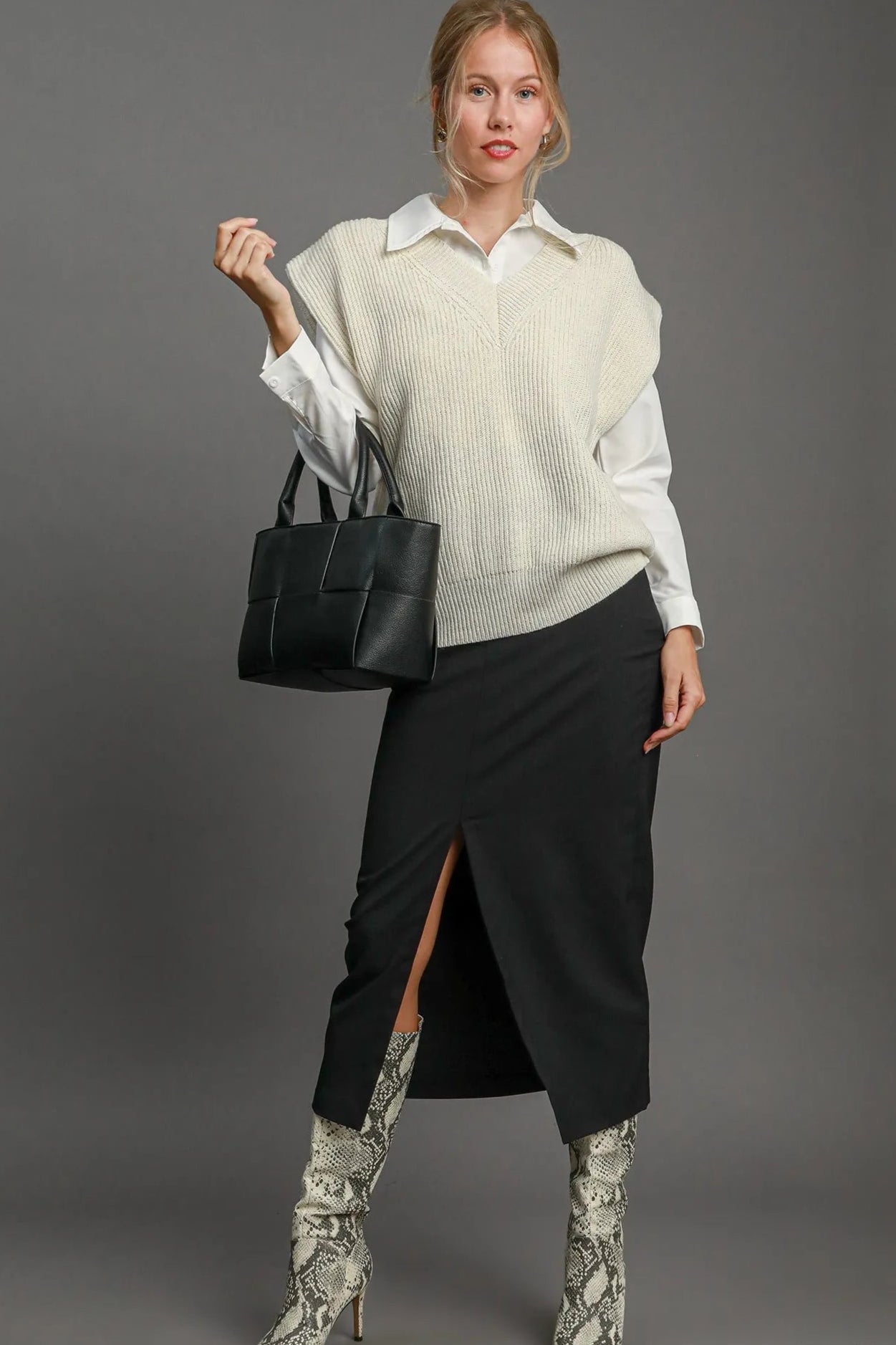 Woman wearing a cream sweater, black skirt, and patterned boots holding a black handbag against a gray background.