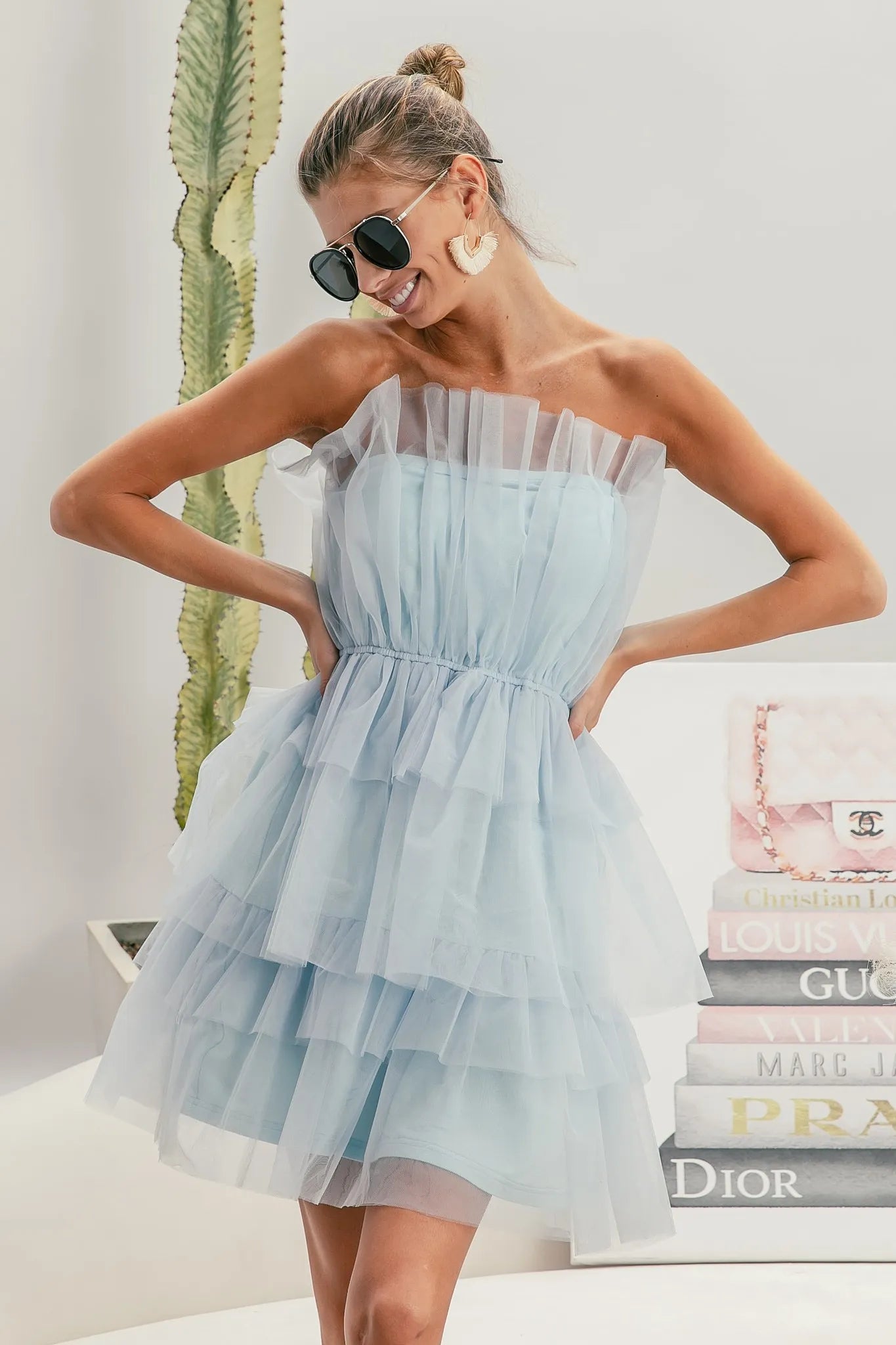 Woman wearing a light blue strapless dress with sunglasses and earrings, standing in front of a stack of fashion magazines.