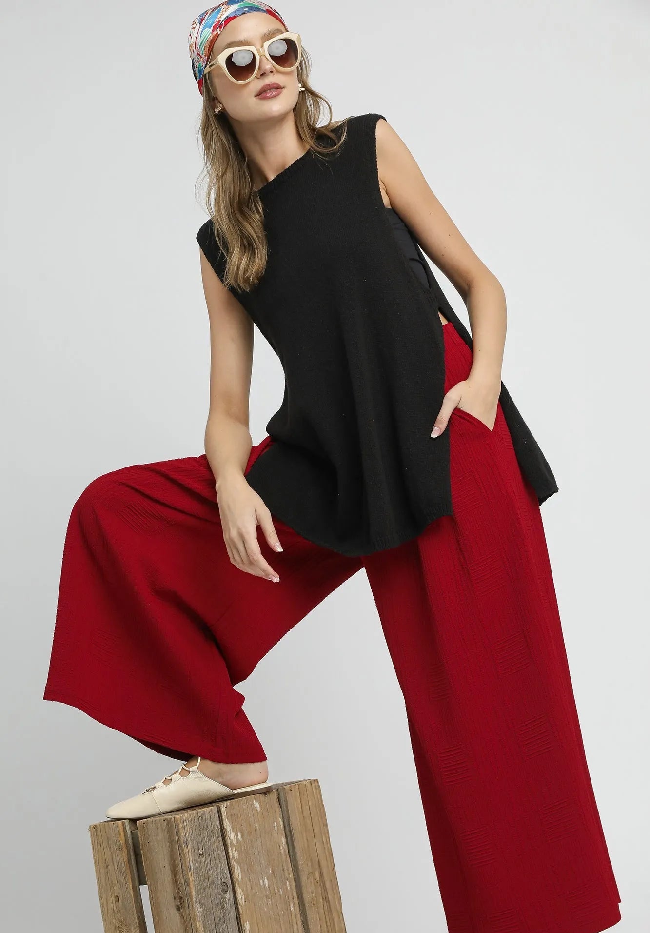 Woman wearing a black sleeveless top and red pants sitting on a wooden block against a gray background.