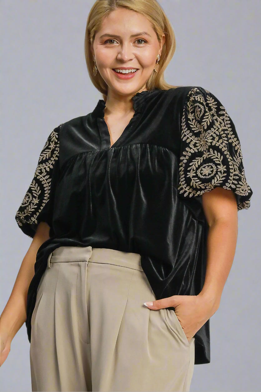 Woman wearing a black blouse with gold embroidery and beige pants on a gray background