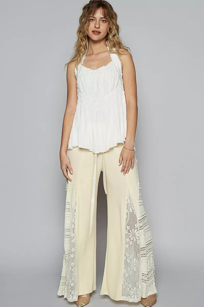 Woman wearing a white sleeveless top and cream pants with lace details on a gray background