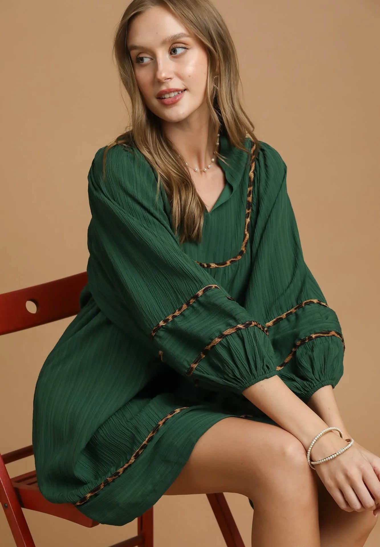Woman wearing a green dress with brown trim sitting on a red chair against a beige background