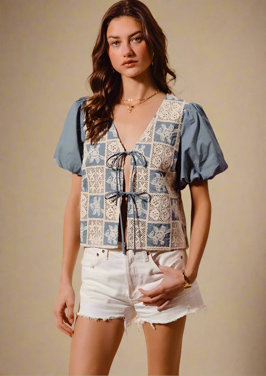 Woman wearing a patterned top with blue sleeves and white shorts against a beige background